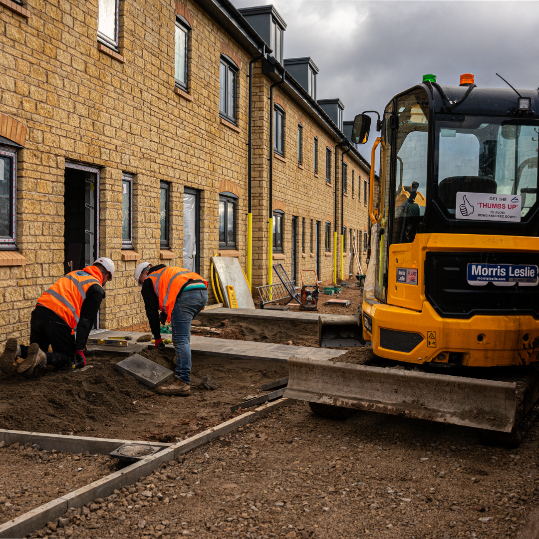 Two SS Construction groundworkers laying paving slabs beside a new build terrace with an excavator on site